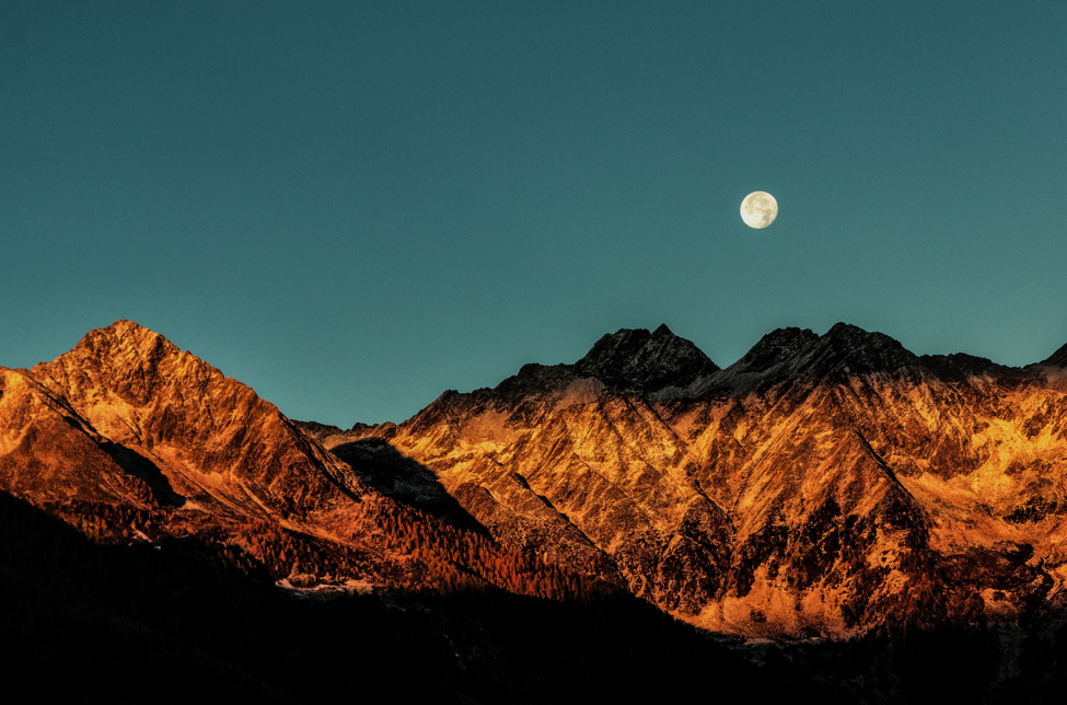 A photo of the moon above golden colored mountains.