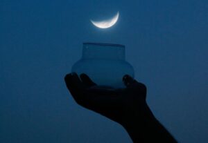 Image of a hand holding a glass jar up to a crescent moon.