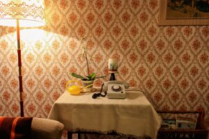 Table with a phone, candle, plant, and watering pot set against a patterned wallpaper. Lamp on the left side of room.